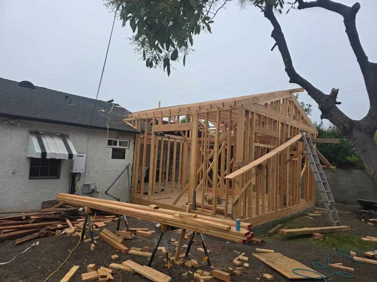 Backyard room addition with gable roof framing by OC Renovation Group in Orange County — wall studs and rafters connecting to existing single-story home